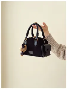 Sac noir en suédine tenu à la main, avec poche latérale, détails dorés et ourson en peluche