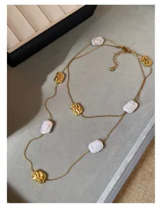 Collana lunga dorata con perle barocche e dettagli quadrati in rilievo, distesa su fondo grigio