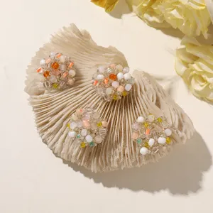 Four colorful beaded stud earrings arranged on a ribbed coral-like prop beside pale yellow flowers.