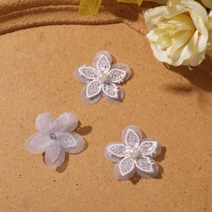 Tre fiori bianchi in pizzo e tulle con centro di perle sono disposti su una superficie color sabbia