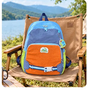 Sac à dos bicolore à panneaux quadrillés et écussons camping, posé sur une chaise au bord du lac
