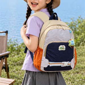 Sac à dos enfant déperlant beige, bleu marine et orange, porté au bord d’un lac en plein air