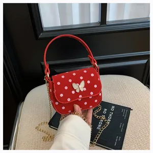 Sac rouge à pois avec fermoir papillon et chaîne dorée, tenu à la main devant un siège beige