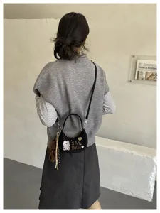 Vue de dos d’un modèle portant un sac noir en velours, brodé «SAY Hello», avec breloque ours