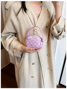 Petit sac rond violet à pois blancs et bordure en dentelle, tenu devant un manteau beige