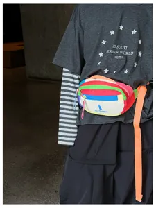 Sac banane à rayures arc-en-ciel, zip bleu et sangle orange porté sur une tenue sombre.