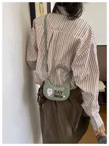 Vue portée d’un sac demi-lune vert, avec broderie d’ours et l’inscription « SAY Hello »