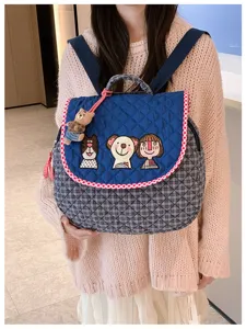 Une personne porte un sac à dos matelassé bleu, avec rabat brodé, bordure à carreaux et ourson.