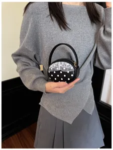 Une main tient un petit sac rond noir à pois blancs et bordure en dentelle devant un pull gris