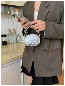 Une main tient un petit sac rond argenté à pois blancs près du téléphone, avec un blazer brun