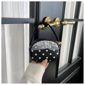 Sac noir arrondi à pois blancs, dentelle et breloque dorée, tenu devant une porte sombre