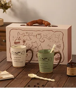 Zwei Tassen mit Katzenmotiv und Keramiklöffel vor einer Geschenkbox auf einem Holztisch