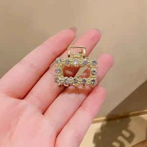Close-up of a square gold-tone clip with clear rhinestones, held in a hand against a beige wall