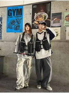 Two models wear black chest bags with adjustable straps and buckles against a wall of posters.