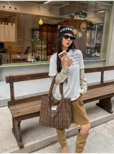 Street-style view of a model carrying a brown plaid tote with long straps beside a wooden bench