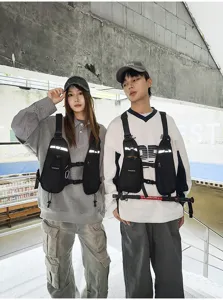Two models wear black chest packs with reflective strips and adjustable straps in an urban setting.