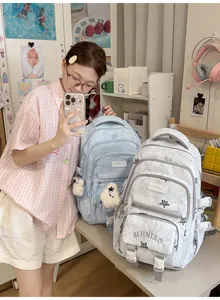 Une fille pose près de sacs à dos bleu et gris clair, avec poches zippées et petits pendentifs ours