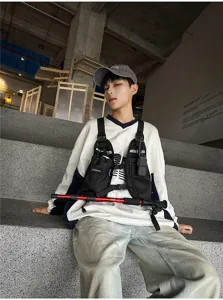 Seated model wearing a black nylon chest vest with utility pockets, webbing straps and buckles