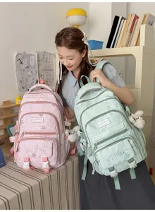Une élève compare deux sacs à dos rose et vert menthe, avec poches avant et oursons en peluche.