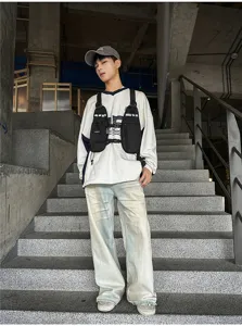 Model wearing a black chest-rig utility bag over a white sweatshirt on outdoor concrete stairs