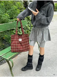 Red plaid fabric tote carried by a model beside a green bench in an outdoor street setting