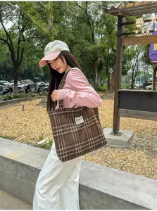 A model carries a brown plaid tote with a white woven label outdoors, shown over her shoulder.