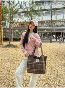 Street-style view of a model carrying a brown plaid canvas tote with double handles outdoors