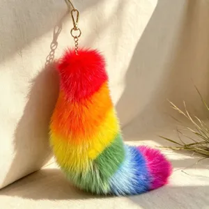 Rainbow faux-fur tail charm with gold-tone clasp and chain, shown on pale fabric in soft light