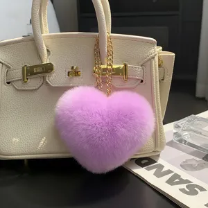 Colgante de corazón mullido en tono lila, con cadena dorada, sobre un bolso color crema