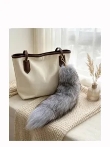 Close-up of a gray fluffy fox-tail charm with a gold clasp hanging from a cream tote bag