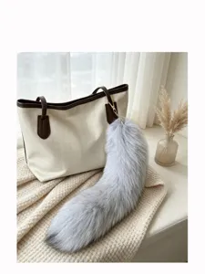 Close-up of a fluffy gray tail charm with a gold clasp hanging from a cream handbag by a window