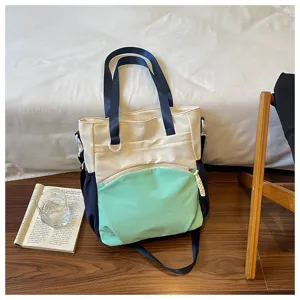 Sac crème, bleu marine et menthe avec poche zippée à l’avant, posé près d’un livre sur un sol en bois.