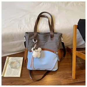 Sac gris et bleu clair, bordure brune et ourson en peluche, posé près de livres et d’une chaise.