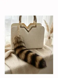 Styled close-up of a fluffy striped tail charm with gold clasp beside an embossed handbag