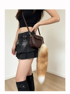 Fluffy orange-brown tail charm with a white tip hangs from a brown shoulder bag in a styled outfit.