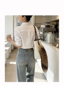 Brown, fluffy fox-tail charm hanging from a cream tote bag, styled with a casual outfit in a café.