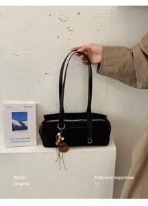 View of a black suede-textured bag with long handles, zipper and wooden charm against a light wall