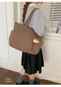 Sac cabas marron porté à l’épaule, avec un pendentif ourson en peluche et des coutures visibles.