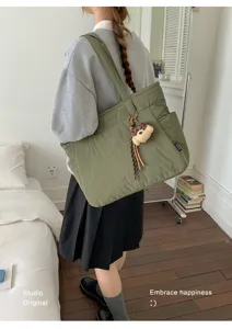 Vue en situation d’un sac vert porté à l’épaule, avec un charm ours en peluche, près d’un lit
