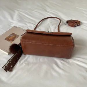 Vista de la base lisa del bolso marrón, con asa superior, correa desmontable y borla con pompón