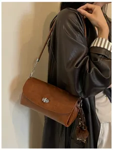 Detalle de un bolso marrón al hombro, con solapa efecto ante, cierre plateado y pompón decorativo