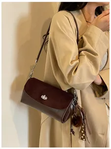 Bolso marrón sobre un abrigo beige, con solapa de ante, cierre giratorio y adorno colgante