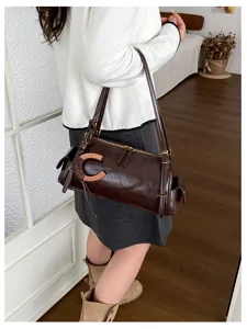 Model wearing a dark brown bag with a long strap, gold zipper, and orange C-shaped ornament