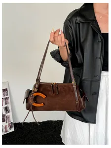 A model holds a brown suede-look bag with a long strap, orange C-shaped accent and tassel charm.