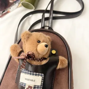 Close-up of a plush brown teddy bear charm with a polka-dot bow on a suede-textured bag