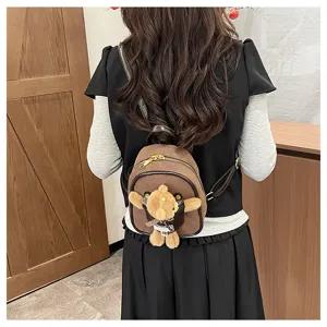 Back view of a model wearing a small brown plush backpack with a teddy bear charm indoors