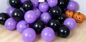 Purple and black balloons with spider motifs arranged on a wooden floor beside glowing pumpkin props