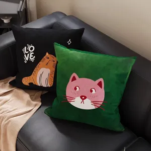 Zwei quadratische Katzenkissen liegen auf einem schwarzen Ledersofa neben einer beigen Decke.