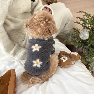 Small dog in a charcoal fuzzy vest with white, orange-centered flowers, seated on a bed