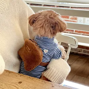Small curly-haired dog wearing a blue-and-white striped turtleneck and a brown plush shoulder bag.